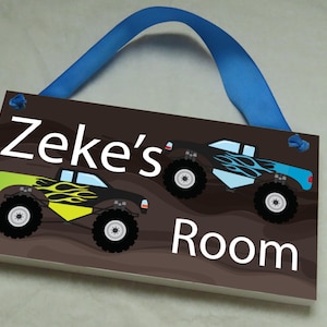 May include: A brown wooden door sign with the text "Zeke's Room" in white. The sign features two cartoon monster trucks with flames and blue and green accents. The sign has a blue ribbon attached for hanging.