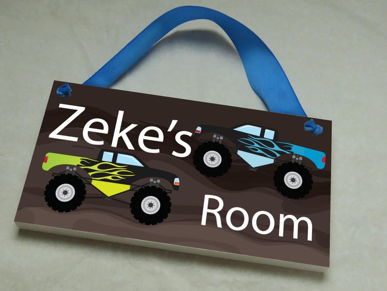 May include: A brown wooden door sign with the text "Zeke's Room" in white. The sign features two cartoon monster trucks with flames and blue and green accents. The sign has a blue ribbon attached for hanging.