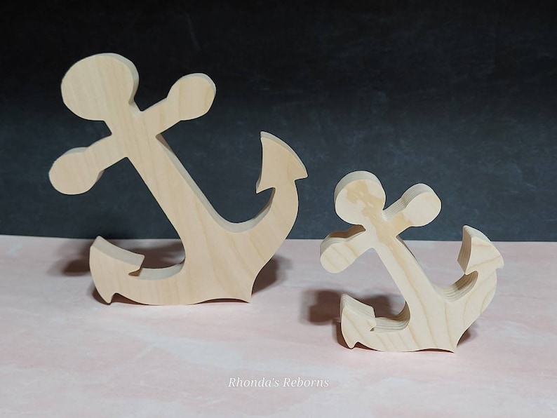 May include: Two wooden anchors, one larger than the other, are shown on a pink surface. Both anchors are made of unfinished wood and have a simple, classic design.