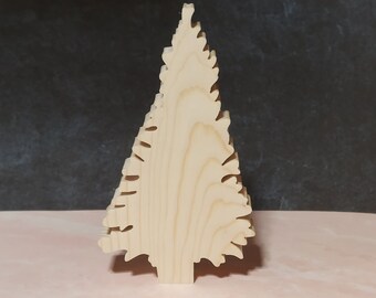 May include: A wooden Christmas tree decoration. The tree is cut from light-colored wood and has a layered design. The edges are cut to resemble the shape of pine needles. The tree stands on a pink surface with a dark gray background.