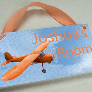 May include: A blue and white wooden sign with a picture of an orange airplane and the text "Joshua's Room". The sign has a light orange ribbon attached to the top for hanging.