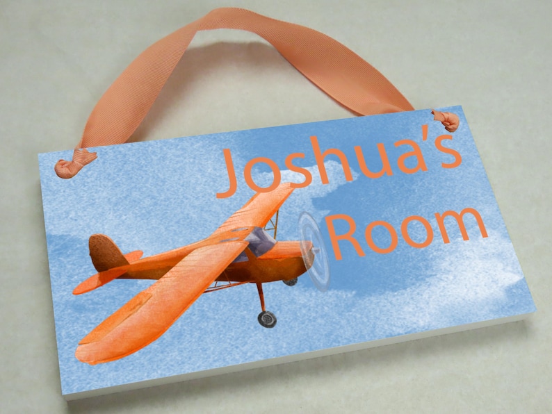 May include: A blue and white wooden sign with a picture of an orange airplane and the text "Joshua's Room". The sign has a light orange ribbon attached to the top for hanging.