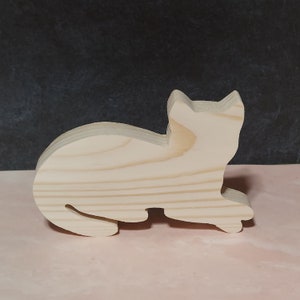 May include: A wooden cutout of a cat in a lying down position. The cat is made of light brown wood and has a smooth, unfinished surface.