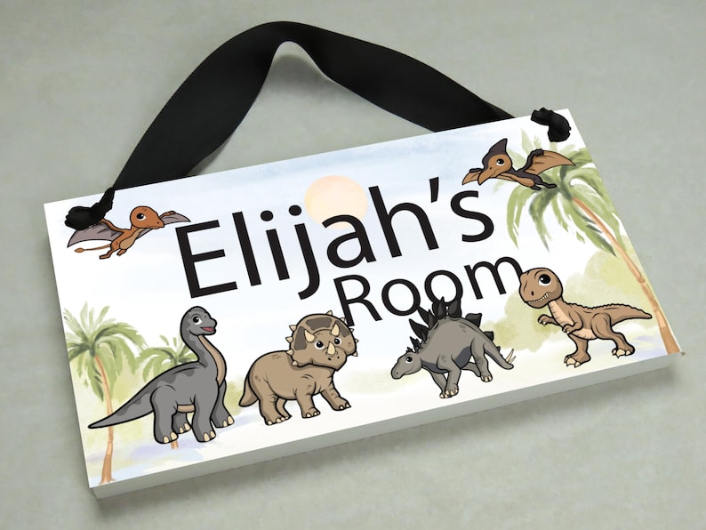 May include: A white wooden door sign with a black ribbon hanger. The sign features a watercolor background with palm trees and dinosaurs. The text "Elijah's Room" is printed in black letters.
