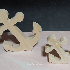 May include: Two wooden anchors, one large and one small, are shown on a pink surface. Both anchors are made of unfinished wood and have a simple, classic design.