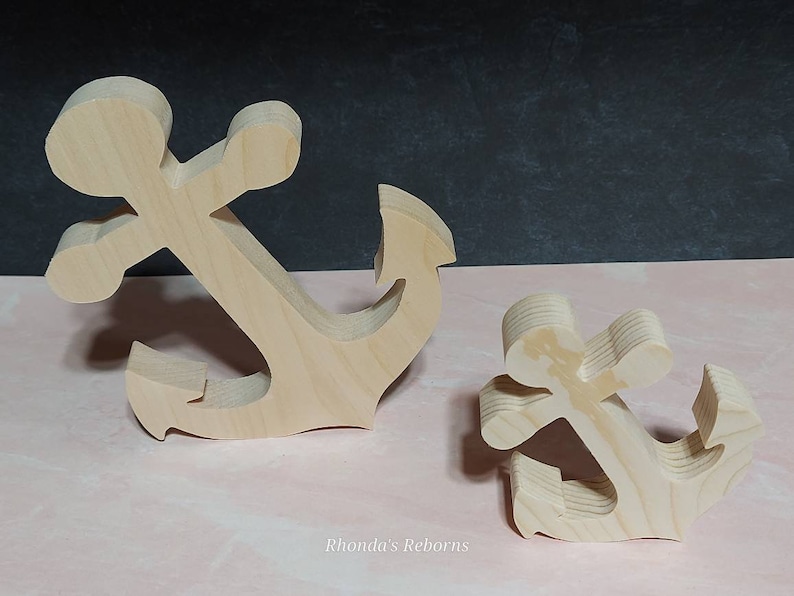 May include: Two wooden anchors, one large and one small, are shown on a pink surface. Both anchors are made of unfinished wood and have a simple, classic design.