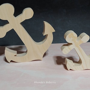 May include: Two wooden anchors, one larger than the other, are displayed on a pink surface. Both anchors are made of unfinished wood and have a simple, classic design.