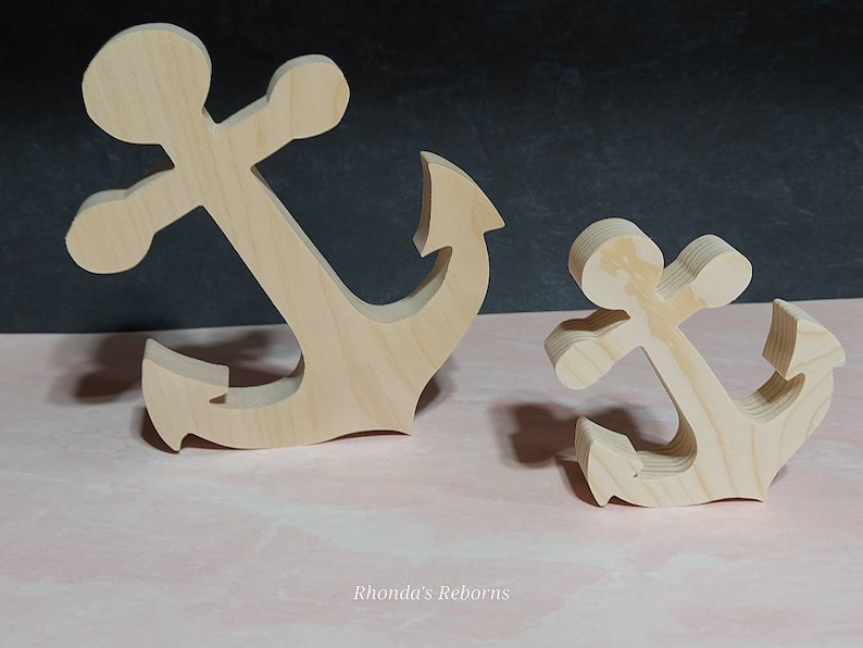 May include: Two wooden anchors, one larger than the other, are displayed on a pink surface. Both anchors are made of unfinished wood and have a simple, classic design.