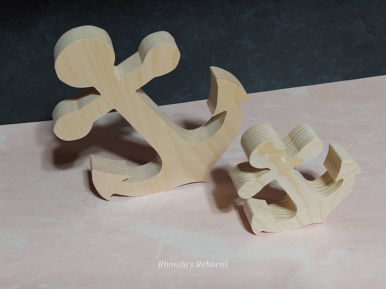 May include: Two wooden anchors, one larger than the other, are shown on a pink surface. The anchors are made of unfinished wood and have a natural grain.