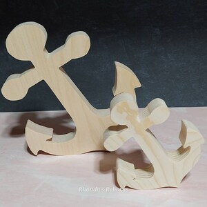 May include: Two wooden anchors, one larger than the other, are displayed on a pink surface. The anchors are unfinished and ready to be painted or stained.