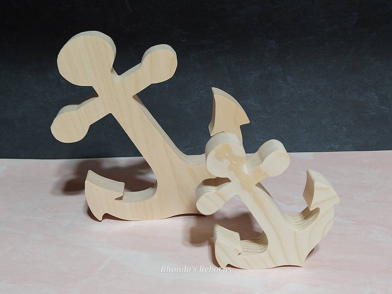 May include: Two wooden anchors, one larger than the other, are displayed on a pink surface. The anchors are unfinished and ready to be painted or stained.
