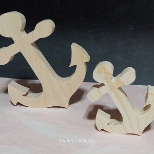 May include: Two wooden anchors, one larger than the other, are shown on a pink surface. Both anchors are made of unfinished wood and have a simple, classic design.