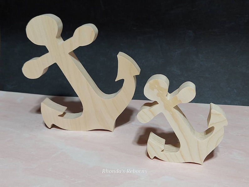 May include: Two wooden anchors, one larger than the other, are shown on a pink surface. Both anchors are made of unfinished wood and have a simple, classic design.
