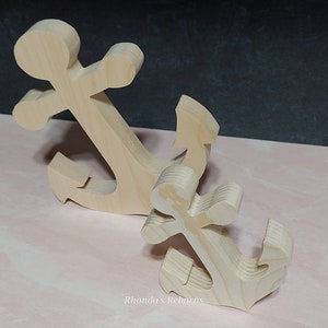 May include: Two wooden anchors, one larger than the other, are shown on a pink surface. The anchors are made of unfinished wood and have a simple design.