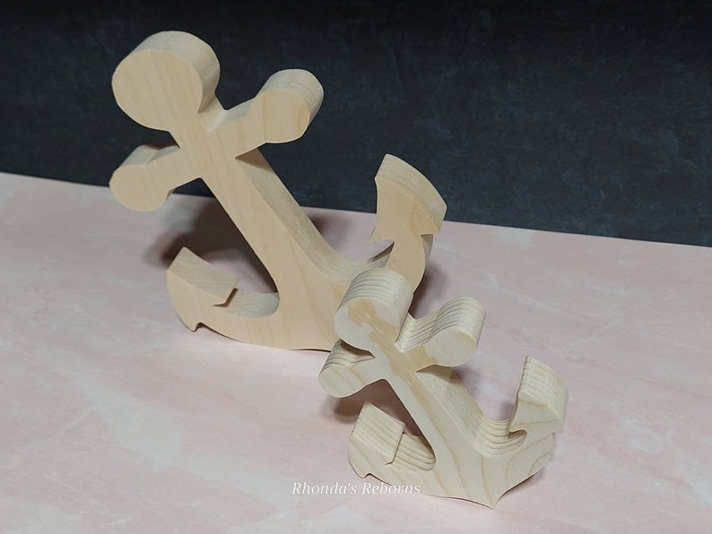 May include: Two wooden anchors, one larger than the other, are shown on a pink surface. The anchors are made of unfinished wood and have a simple design.