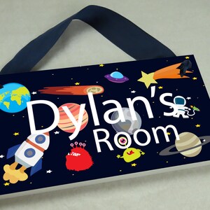 May include: A dark blue wooden sign with a space theme. The sign has a white background with the text "Dylan's Room" in white letters. The sign is decorated with planets, stars, a rocket, and an alien.