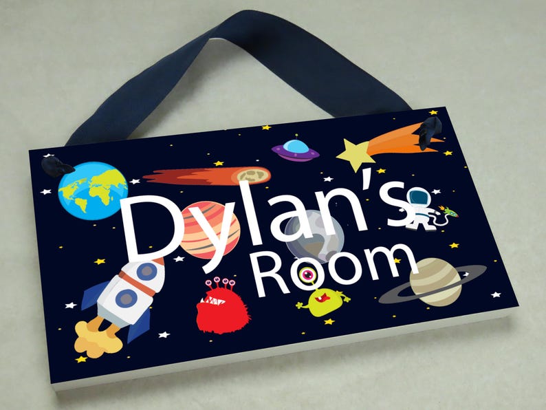 May include: A dark blue wooden sign with a space theme. The sign has a white background with the text "Dylan's Room" in white letters. The sign is decorated with planets, stars, a rocket, and an alien.