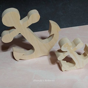 May include: Two wooden anchors, one larger than the other, are shown on a pink surface. The anchors are made of unfinished wood and have a natural grain.