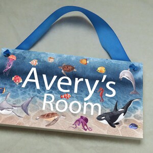 May include: A blue and white wooden door sign with a watercolor ocean scene. The sign says "Avery's Room" and features sea creatures like a shark, dolphin, seahorse, and octopus.