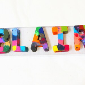 May include: A name spelled out in colorful crayon letters, the letters are "BLAIR" and are arranged in a row on a white surface.