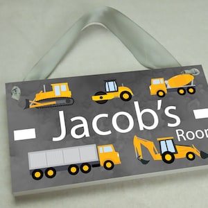 May include: A gray wooden sign with a white border and the text "Jacob's Room" in white. The sign is decorated with yellow construction vehicles, including a bulldozer, a steamroller, a cement mixer, a dump truck, and an excavator.