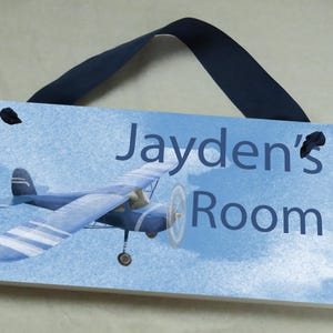May include: A blue and white airplane with a blue ribbon hanging from the top. The sign reads "Jayden's Room" on a blue background with white clouds.