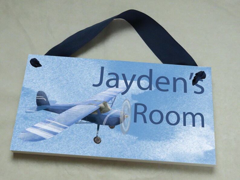 May include: A blue and white airplane with a blue ribbon hanging from the top. The sign reads "Jayden's Room" on a blue background with white clouds.