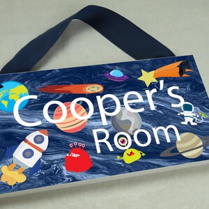 May include: A blue and white wooden door sign with the text "Cooper's Room" in white. The sign features a space theme with planets, stars, a rocket, and aliens.