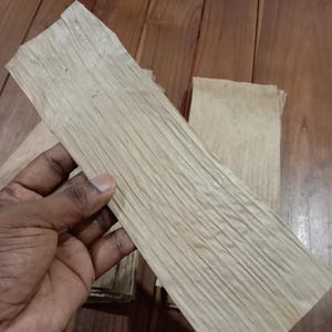 May include: A stack of light brown, textured wooden sheets. The sheets have a natural, organic appearance with visible grain patterns. The wood is held by a hand, with other sheets visible in the background. The sheets appear to be approximately 12 inches long.