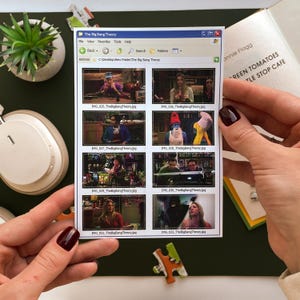 May include: A sticker featuring a Windows-style interface with images from "The Big Bang Theory" TV show. The sticker includes the show's title and file names. A small potted plant, headphones, and a book are also visible.