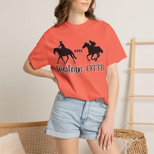 May include: A coral-colored short-sleeve t-shirt featuring a black graphic of two horses with riders and the text Western OTTB. The shirt has a crew neckline and is paired with light blue denim shorts that have a cuffed hem at the bottom.