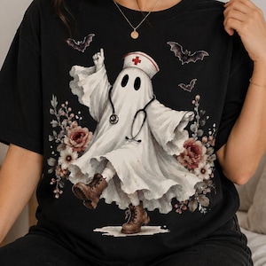 May include: A black t-shirt featuring a graphic of a white ghost dressed as a nurse, wearing a nurse cap with a red cross and a stethoscope. The ghost is surrounded by floral accents and flying bats, and is wearing brown lace-up boots.