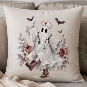 May include: A square throw pillow featuring a white ghost wearing a nurse cap with a red cross, a stethoscope, and brown lace-up boots. The ghost is surrounded by watercolor-style red and white flowers and three small black flying bats.