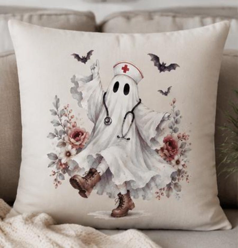 May include: A square throw pillow featuring a white ghost wearing a nurse cap with a red cross, a stethoscope, and brown lace-up boots. The ghost is surrounded by watercolor-style red and white flowers and three small black flying bats.