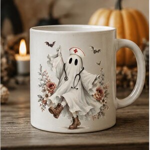 May include: A white ceramic mug featuring a graphic of a ghost dressed as a nurse, complete with a stethoscope and a nurse cap with a red cross. The ghost is surrounded by floral accents and small bats, wearing brown lace-up boots.
