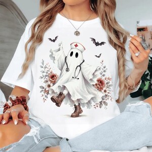 May include: A white t-shirt featuring a graphic of a ghost dressed as a nurse, wearing a nurse cap with a red cross and a stethoscope. The ghost is surrounded by floral accents and flying bats, and is wearing brown lace-up boots.
