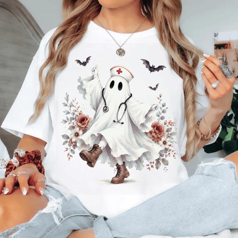 May include: A white t-shirt featuring a graphic of a ghost dressed as a nurse, wearing a nurse cap with a red cross and a stethoscope. The ghost is surrounded by floral accents and flying bats, and is wearing brown lace-up boots.
