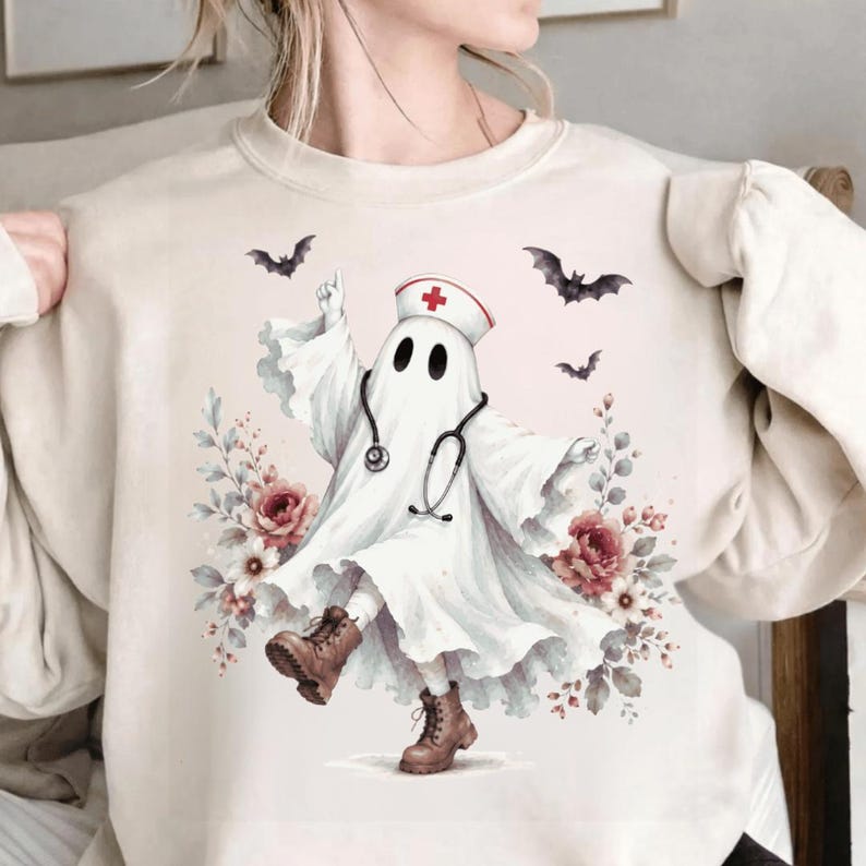 May include: A cream-colored crewneck sweatshirt featuring a graphic of a ghost dressed as a nurse, wearing a white cap with a red cross and a stethoscope. The ghost is surrounded by pink flowers, green leaves, and black bats in flight.