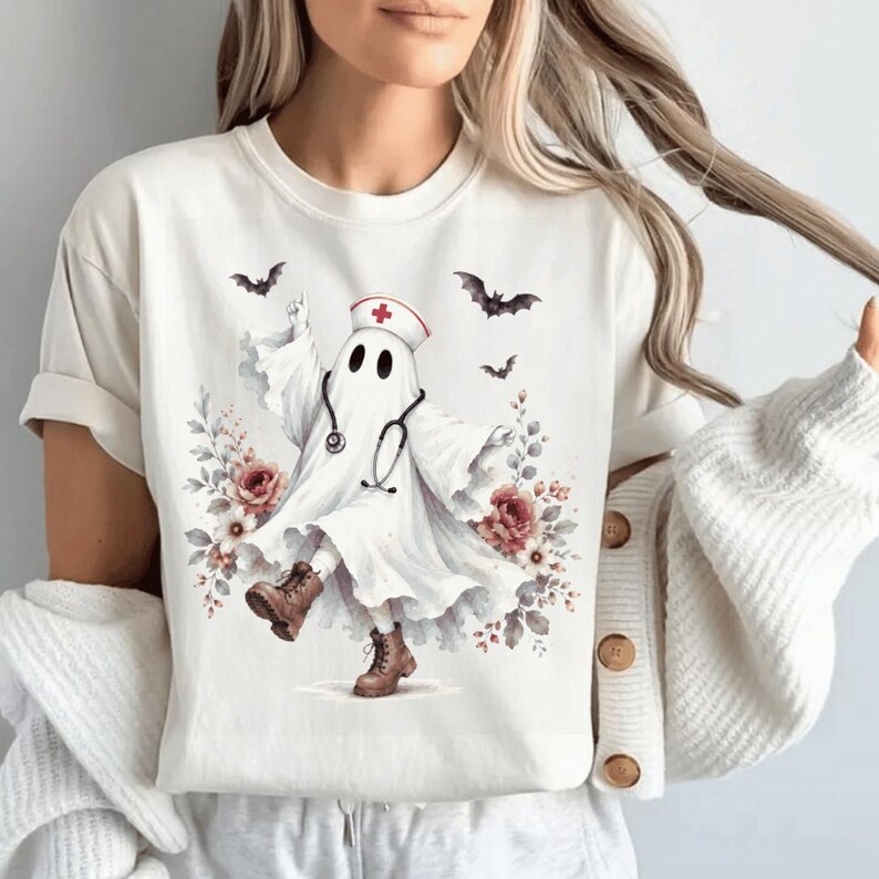 May include: A cream-colored short-sleeve t-shirt featuring a graphic of a ghost wearing a nurse cap with a red cross, a stethoscope, and brown boots. The ghost is surrounded by floral arrangements in shades of pink and red, with small black bats flying above.