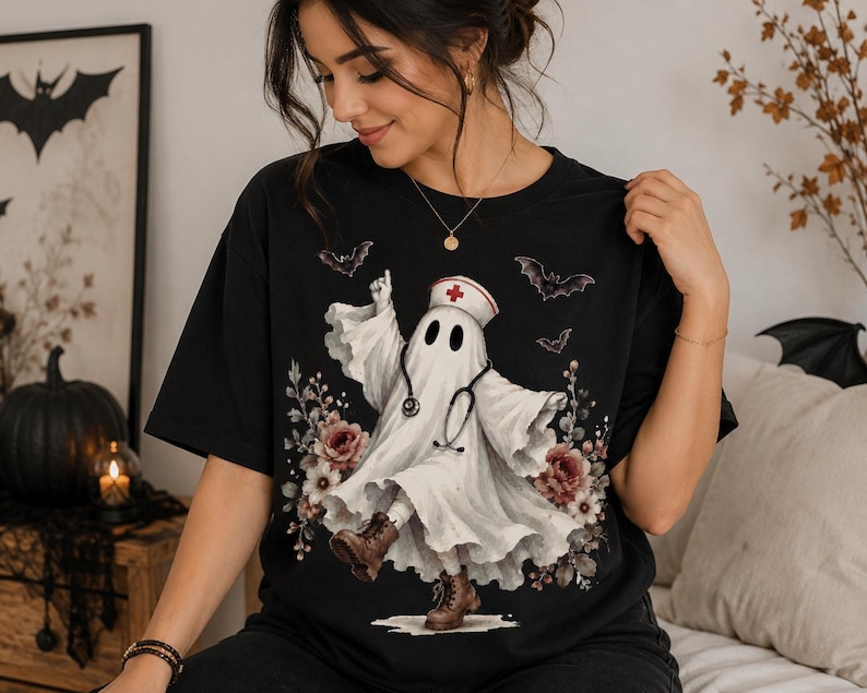 May include: A black t-shirt featuring a graphic of a white ghost dressed as a nurse, wearing a nurse cap with a red cross and a stethoscope. The ghost is surrounded by floral accents and flying bats, and is wearing brown lace-up boots.