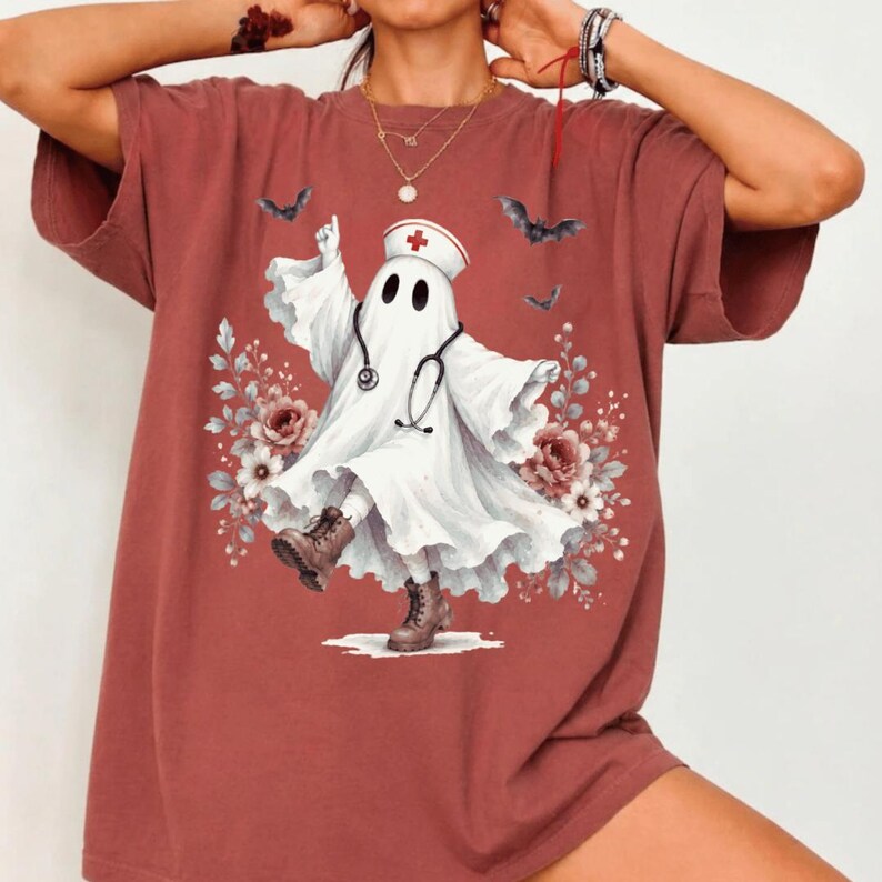 May include: A brick-red short-sleeve t-shirt featuring a graphic of a white ghost dressed as a nurse, wearing a cap with a red cross and a stethoscope. The ghost is surrounded by floral accents and small black bats, wearing brown boots.