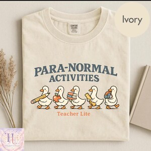 Puede incluir: Una camiseta de color marfil con el texto PARA-NORMAL ACTIVITIES en azul oscuro, con cinco patos blancos debajo, cada uno cargando útiles escolares como un lápiz, manzana, libros, regla y mochila, con el texto Teacher Lite en naranja debajo.