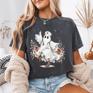 May include: A charcoal gray short-sleeve t-shirt featuring a graphic of a white ghost dressed as a nurse with a stethoscope and cap. The ghost is surrounded by floral accents and small bats, with a vintage-style wash on the fabric.