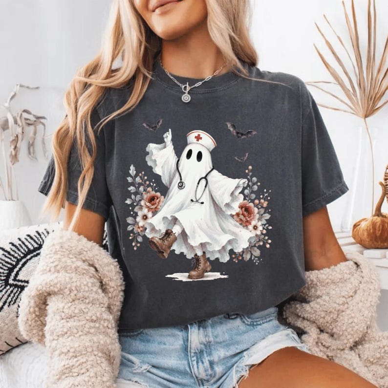 May include: A charcoal gray short-sleeve t-shirt featuring a graphic of a white ghost dressed as a nurse with a stethoscope and cap. The ghost is surrounded by floral accents and small bats, with a vintage-style wash on the fabric.