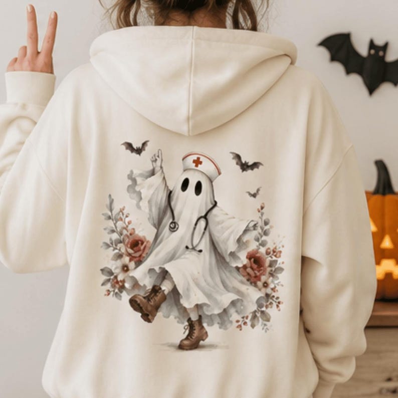 May include: A cream-colored hooded sweatshirt featuring a graphic on the back of a ghost dressed as a nurse with a stethoscope, wearing brown boots, surrounded by red roses and small black bats flying in the air against a light background.