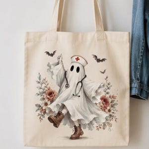 May include: A beige canvas tote bag featuring a graphic of a ghost dressed as a nurse, wearing a white cap with a red cross, a stethoscope, and brown boots. The ghost is surrounded by floral arrangements and small flying bats in a soft color palette.