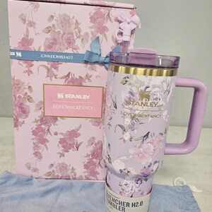 May include: A floral-patterned Stanley Quencher H2.0 tumbler and matching gift box. The tumbler is lavender with a handle and a floral design. The gift box is pink with a floral pattern and a blue ribbon. The text "LOVESHACKFANCY" and "STANLEY" are visible.