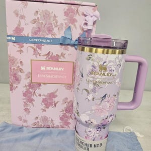 May include: A lavender Stanley Quencher H2.0 tumbler with a floral design, a matching handle, and a lid. The tumbler is next to a gift box with a similar floral pattern and a blue ribbon. The box and tumbler have the "LOVESHACKFANCY" and "STANLEY" logos.