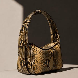 Puede incluir: Un bolso de estilo hobo estructurado con un patrón de piel de serpiente en amarillo y negro. El bolso cuenta con una sola correa de hombro integrada, una abertura superior curva y ribetes negros a lo largo de los bordes, creando una silueta definida.
