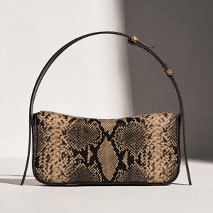 Può includere: Una borsa a spalla rettangolare con un motivo in pelle di serpente color beige e nero. La borsa presenta una silhouette sottile e curva e un cinturino in pelle nera sottile e regolabile con piccoli dettagli metallici sui lati.